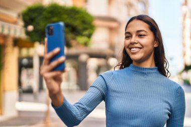 Genç Afrikalı Amerikalı kadın gülümsüyor. Kendine güveni tam. Sokakta akıllı telefondan selfie çekiyor.