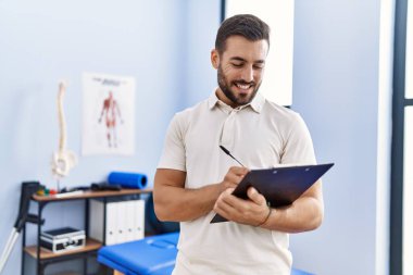 Fizyoterapist üniforması giyen genç İspanyol adam klinikteki panoya yazı yazıyor.