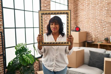 Hispanic woman at new home holding empty frame clueless and confused expression. doubt concept. 