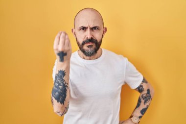 Sarı arka planda duran İspanyol dövmeli genç adam. İtalyan jesti yapıyor. Elleri ve parmakları kendinden emin. 