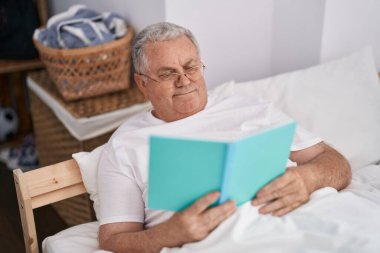 Orta yaşlı, kır saçlı, kitap okuyan, yatak odasında oturan bir adam.