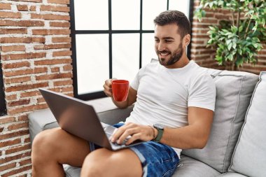 Genç İspanyol adam dizüstü bilgisayar kullanıyor ve evde kahve içiyor.