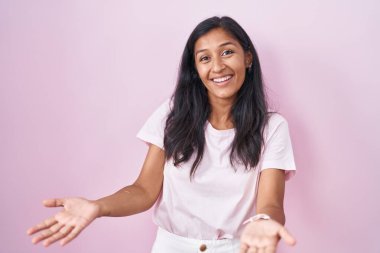 Young hispanic woman standing over pink background smiling cheerful with open arms as friendly welcome, positive and confident greetings 