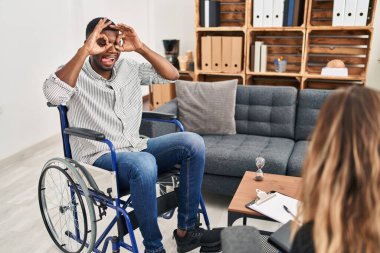 African american man doing therapy sitting on wheelchair doing ok gesture like binoculars sticking tongue out, eyes looking through fingers. crazy expression. 