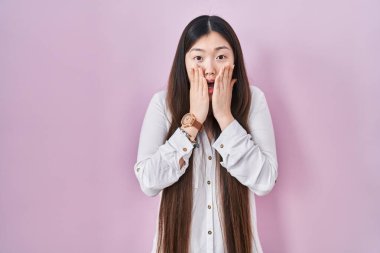 Chinese young woman standing over pink background afraid and shocked, surprise and amazed expression with hands on face 