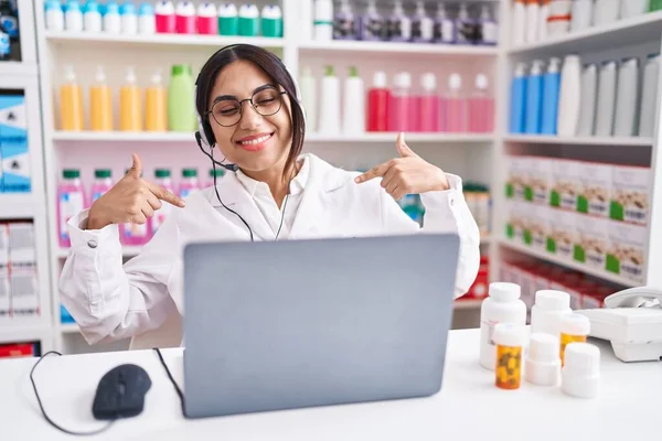Young arab woman working at pharmacy drugstore using laptop looking confident with smile on face, pointing oneself with fingers proud and happy.
