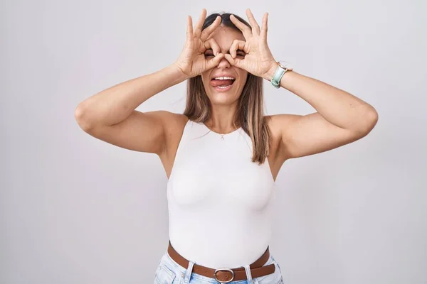 白い背景の上に立つヒスパニック系の若い女性は 双眼鏡の舌を突き出すような をジェスチャーを行うと 目の指を見て クレイジーな表現 — ストック写真