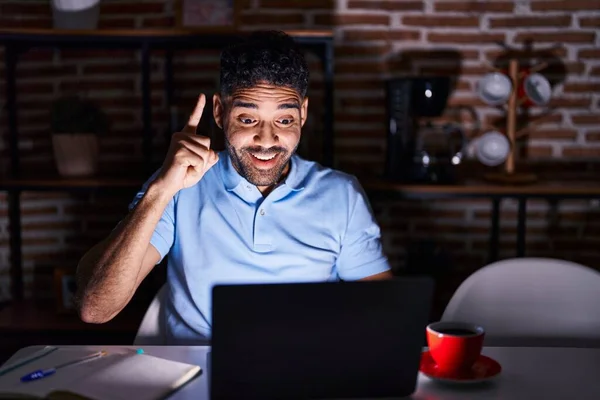 Hispanic man with beard using laptop at night pointing finger up with successful idea. exited and happy. number one.
