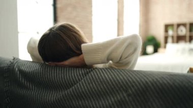 Sarışın genç kadın ellerini başının üstüne koyup evdeki kanepede oturuyor.