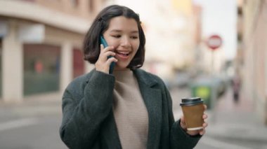 Young hispanic girl talking on the smartphone drinking coffee at street