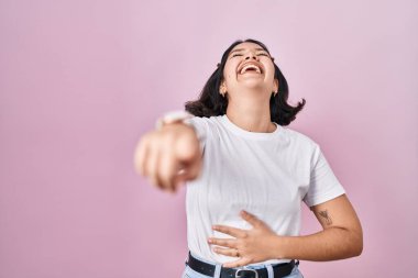 Pembe arka plan üzerine günlük beyaz t-shirt giyen genç İspanyol kadın sana gülüyor, eli vücudunun üzerinde kamerayı işaret ediyor, utanç ifadesi. 
