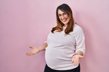 Pregnant woman standing over pink background smiling cheerful with open arms as friendly welcome, positive and confident greetings 