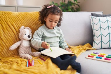 Adorable hispanic girl drawing on notebook sitting on sofa at home