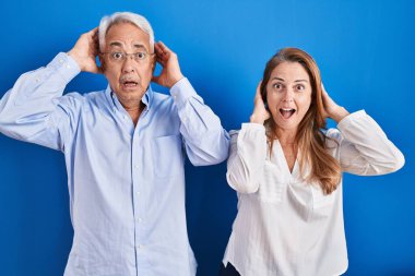 Middle age hispanic couple standing over blue background crazy and scared with hands on head, afraid and surprised of shock with open mouth 