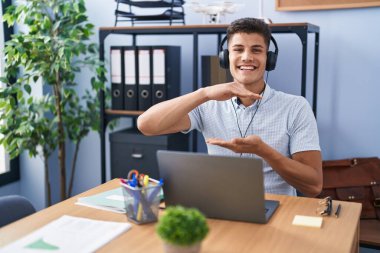 Young hispanic man working at the office wearing headphones gesturing with hands showing big and large size sign, measure symbol. smiling looking at the camera. measuring concept. 