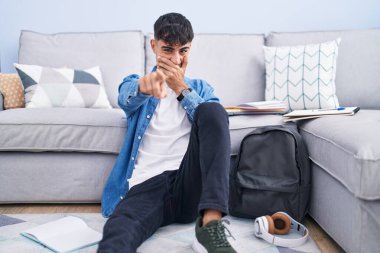 Young hispanic man sitting on the floor studying for university laughing at you, pointing finger to the camera with hand over mouth, shame expression 