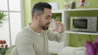 Young hispanic man drinking water sitting on table at home