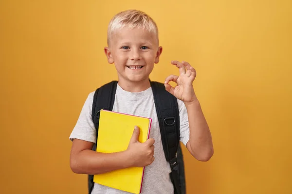 Little caucasian boy wearing student backpack and holding book doing ok sign with fingers, smiling friendly gesturing excellent symbol
