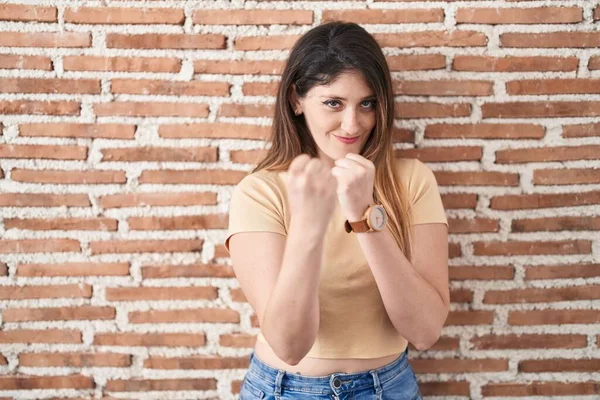 Young brunette woman standing over bricks wall ready to fight with fist defense gesture, angry and upset face, afraid of problem