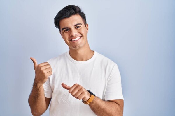 Hispanic man standing over blue background pointing to the back behind with hand and thumbs up, smiling confident 