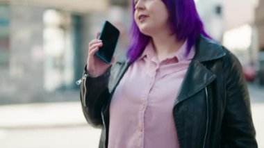 Genç ve cüsseli bir kadın gülümseyerek sokakta akıllı telefondan konuşuyor.