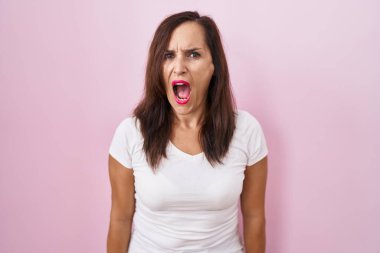 Middle age brunette woman standing over pink background angry and mad screaming frustrated and furious, shouting with anger. rage and aggressive concept. 