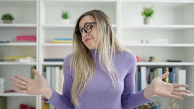 Young blonde woman student standing with i dont know expression at library university