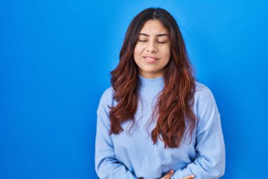 Hispanic young woman standing over blue background with hand on stomach because indigestion, painful illness feeling unwell. ache concept. 