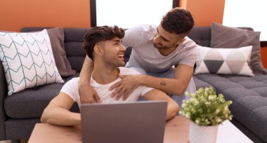 Two hispanic men couple using laptop sitting on sofa at home
