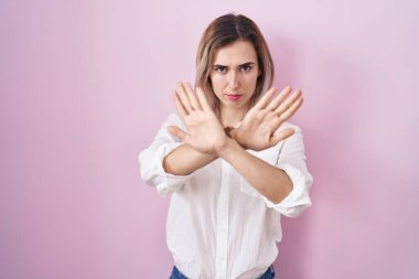 Young beautiful woman standing over pink background rejection expression crossing arms and palms doing negative sign, angry face 