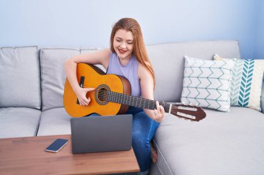 Genç kızıl saçlı kadın evdeki kanepede oturmuş online klasik gitar dersi alıyor.