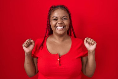 African american woman with braided hair standing over red background celebrating surprised and amazed for success with arms raised and open eyes. winner concept. 