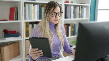 Young blonde woman student using computer and touchpad studying at library university