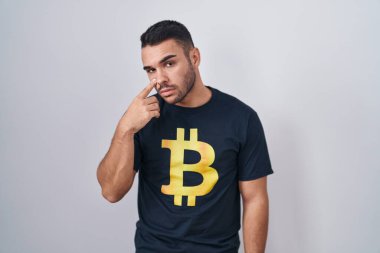 Young hispanic man wearing bitcoin t shirt pointing to the eye watching you gesture, suspicious expression 