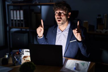 Hispanic young man working at the office at night amazed and surprised looking up and pointing with fingers and raised arms. 