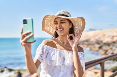 Young brazilian woman taking a selfie photo with smartphone outdoors doing ok sign with fingers, smiling friendly gesturing excellent symbol 