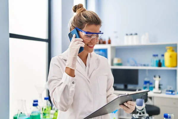 若いですブロンド女性身に着けています科学者制服を着て話す上のスマートフォンで実験室 — ストック写真
