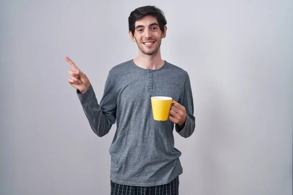 Young hispanic man wearing pajama drinking a cup of coffee with a big smile on face, pointing with hand finger to the side looking at the camera.
