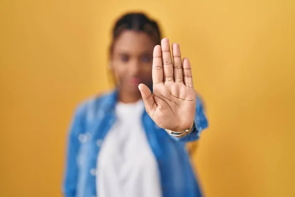 African american woman with braids standing over yellow background doing stop sing with palm of the hand. warning expression with negative and serious gesture on the face.