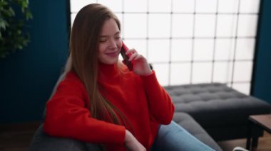 Genç sarışın kadın akıllı telefonda konuşuyor. Evdeki kanepede oturuyor.