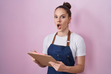 Young hispanic girl wearing professional waitress apron taking order afraid and shocked with surprise and amazed expression, fear and excited face. 