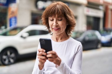 Sokakta akıllı telefon kullanan orta yaşlı bir kadın gülümsüyor.