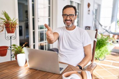 Middle age man using computer laptop at home smiling friendly offering handshake as greeting and welcoming. successful business. 