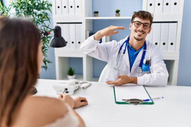 Young doctor with client at medical clinic gesturing with hands showing big and large size sign, measure symbol. smiling looking at the camera. measuring concept. 