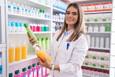 Young beautiful hispanic woman pharmacist holding shampoo bottles at pharmacy