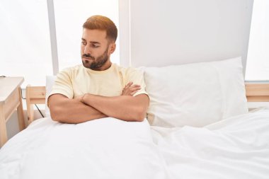 Young hispanic man sitting on bed with unhappy expression and arms crossed gesture at bedroom