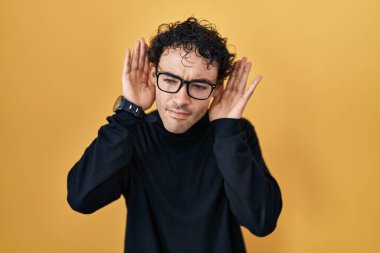 Hispanic man standing over yellow background trying to hear both hands on ear gesture, curious for gossip. hearing problem, deaf 