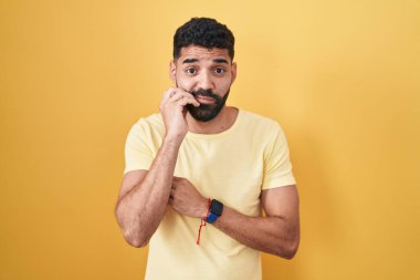 Hispanic man with beard standing over yellow background looking stressed and nervous with hands on mouth biting nails. anxiety problem. 