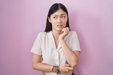 Pembe arka planda duran Çinli genç bir kadın gergin ve gergin görünüyor. Elleri ağızdan tırnak yiyor. anksiyete. 