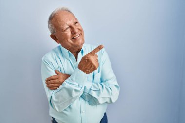 Senior man with grey hair standing over blue background with a big smile on face, pointing with hand and finger to the side looking at the camera. 